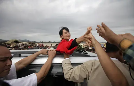 Aung San Suu Kyi reaches out of the top of a car and holds a supporter's hand.