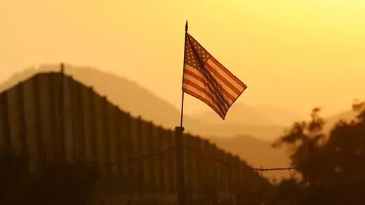 The U.S. flag appears against a yellow sky with a dark fence looming behind it.