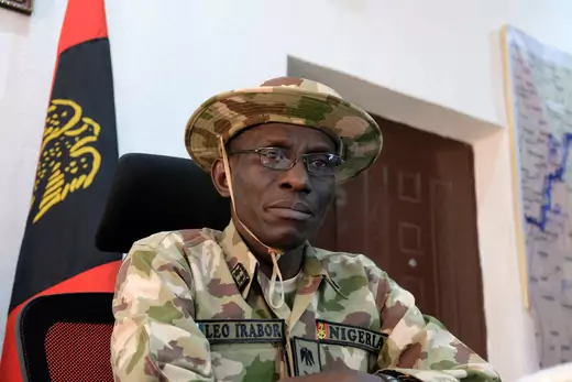 Major General Leo Irabor, then-commander of the Nigerian military's operation against Islamist insurgency Boko Haram, is seated while speaking to the media. He is wearing camouflage, military fatigues with a hat. A flag is on a flagpole over his right shoulder.