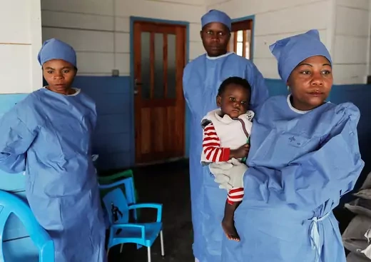 Two men and one woman are seen wearing blue scrubs at an Ebola treatment center. The woman is holding a baby.