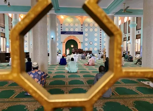 Several Muslim worshippers are seen from from behind sitting on the floor inside a mosque. They are socially distanced, as suggested by health protocols for COVID-19. Most are wearing Islamic dress.
