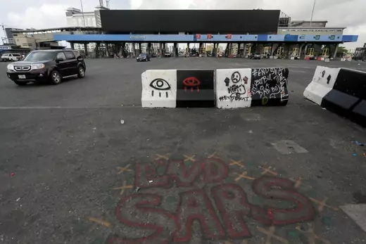 A picture of the Lekki toll gate, with concrete road barriers and a passing car in the foreground. Graffiti is sprayed on the barriers, with protest slogans such as #EndSARS written in chalk on the pavement.