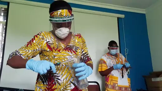 Two women in Tanzania are seen wearing gloves, masks, and facial shields to protect against COVID-19. Both are also wearing colorful, patterned clothing.