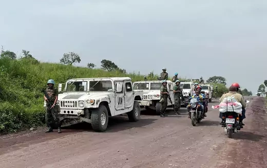 Four UN vehicles are seen parked on a dirt road, protected by armed UN soldiers, in eastern DRC. Two motorcycles are also seen driving on the road, going in opposite directions.