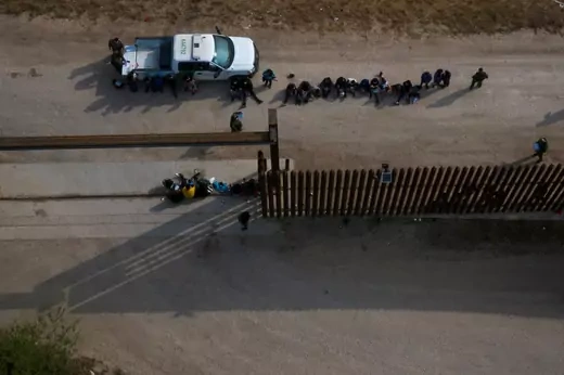 An aerial photo of a line of people sitting near a white pickup truck. They are facing a border barrier.