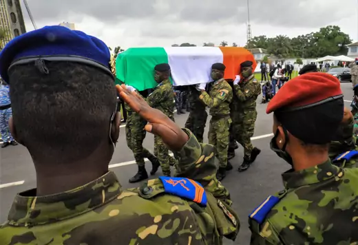 Two soldiers are seen from the rear saluting a coffin of a soldier, with the Ivory Coast flag draped over top, being carried by several members of Ivory Coast's military.