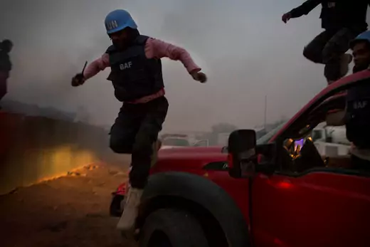 A UN peacekeeper wearing a blue helmet jumps from a red truck while a fire burns behind him.