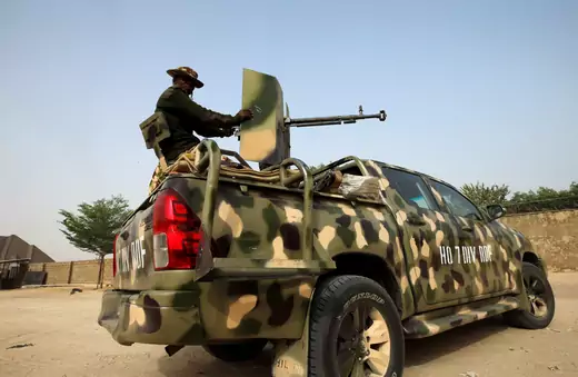 A camouflaged Nigerian military truck with a soldier sitting in the back manning a mounted gun.