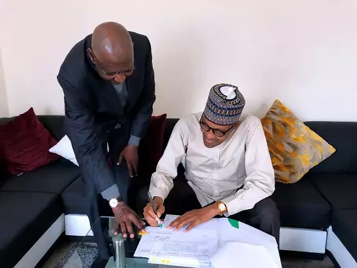 Nigerian President Muhammadu Buhari is seated, wearing traditional dress, while signing a bill into law.