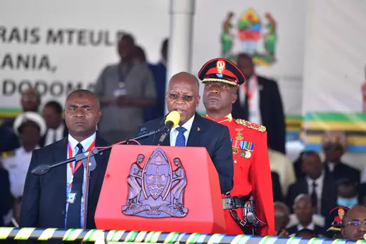 Tanzanian President John Magufuli, standing at a red podium with his country's coat of arms, gives a speech after re-election. He is flanked by a soldier in a red uniform and one other official wearing professional dress.