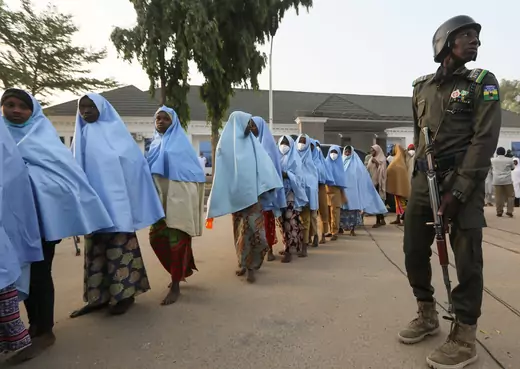 A line of young girls, most without shoes and wearing blue Islamic headdress, leave a building after being released from captivity by bandits. A Nigerian policeman carrying an assault rifle is standing next to the line of girls.