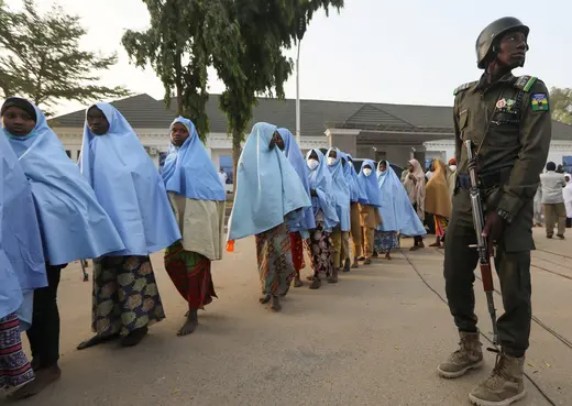 A line of young girls, most without shoes and wearing blue Islamic headdress, leave a building after being released from captivity by bandits. A Nigerian policeman carrying an assault rifle is standing next to the line of girls.