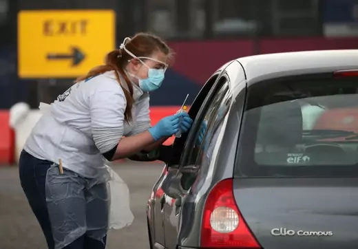 At a COVID-19 outdoor testing site a woman in PPE uses a nasal swab to test a passenger in a car for COVID-19. 