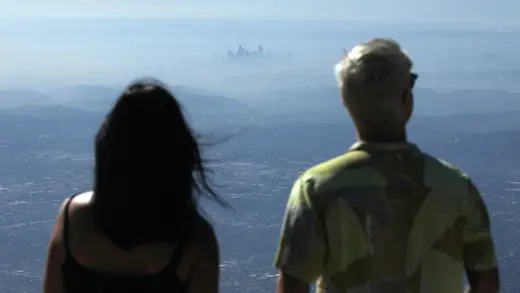 Two people look out over downtown Los Angeles, which appears in a cloud of haze.