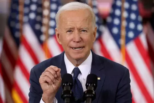 Close-up of Joe Biden in a blue suit and blue tie, speaking and gesturing with his right hand, in front of several U.S. flags