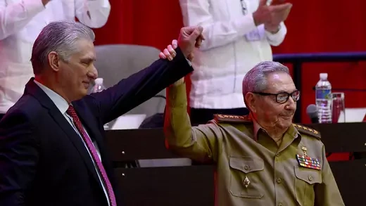 Dressed in an olive green military outfit, Raul Castro holds up the hand of Miguel Diaz-Canel, who is wearing a suit. People in the background are applauding.