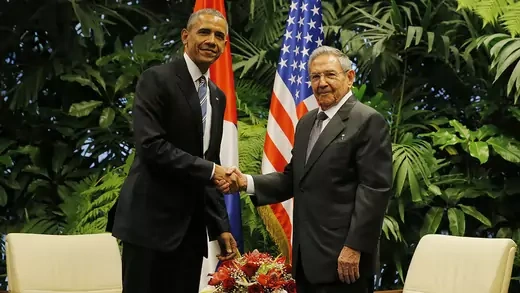 U.S. President Barack Obama and Cuban President Raul Castro shake hands.  Tropical plants, flags, and two chairs appear behind them.