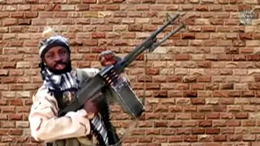 Abubakar Shekau, the leader of Boko Haram, stands in front of a brick wall holding a heavy machine gun.