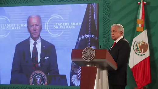 Mexico's President Andres Manuel Lopez Obrador stands at a podium next to U.S. President Joe Biden who is on a large screen speaking virtually.
