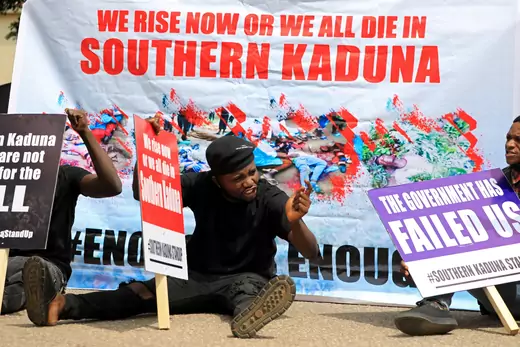 A man sits on the ground in front of a sign that says "We rise now or we all die in Southern Kaduna." Another sign says "The government has failed us."