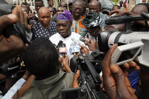 Former Nigerian President Olusegun Obasanjo is seen with a large group of reporters holding cameras and microphones.