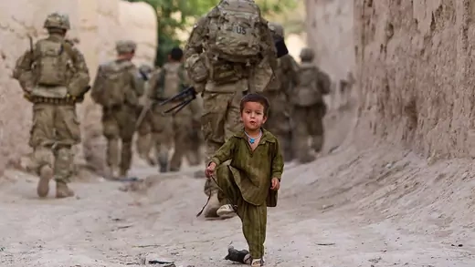 A boy plays on a street as several U.S. Army soldiers walk away on patrol in the background