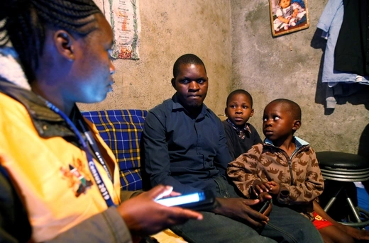 A man and his two children sit and speak to a woman wearing a bright orange vest and holding a smartphone who is working as an enumerator for the Kenyan Census.