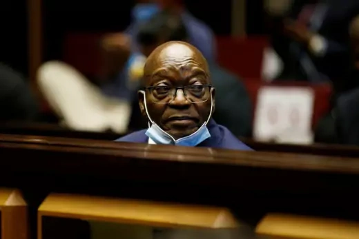 A picture of former South African President Jacob Zuma sitting in a courtroom during a trial on his involvement in corruption. He is wearing a facemask, situated on his chin, due to the country's ongoing COVID-19 outbreak.