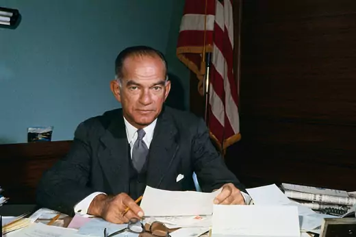 Senator J. William Fulbright sits behind his desk holding papers and looking at the camera. 