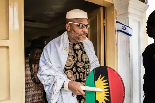 Nnamdi Kanu, leader of the Indigenous People of Biafra, emerges from a room wearing patterned clothing and holding a shield with the flag of Biafra, a breakaway state defeated in Nigeria's civil war in 1967-70. A flag of Israel is also next to the doorway, signifying Kanu's Jewish faith.