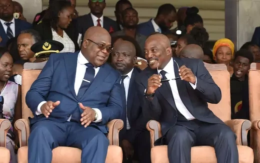 Current President of the Democratic Republic of Congo Felix Tshisekedi and his predecessor Joseph Kabila sit on plush orange seats while wearing suits in apparent conversation.