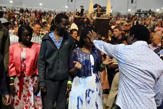 T.B. Joshua, a now-deceased Nigerian pastor, holds his hand on a woman's face. The woman holds her hands raised with her palms upward.