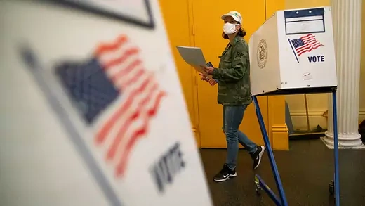 Voting booth with U.S. flag in the forefront as a woman walks with her ballot in the background