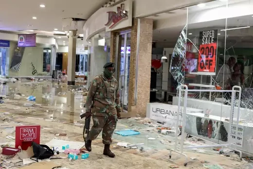 A South African soldier walks through a looted mall with an assault rifle in hand. Shards of broken glass litter the area under his feet.