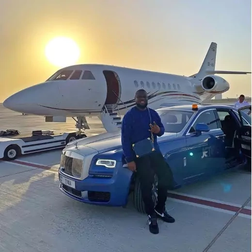 A man, with a designer bag, leans against the hood of a blue Rolls Royce with a private jet in the background. The sun is seen setting above the plane.