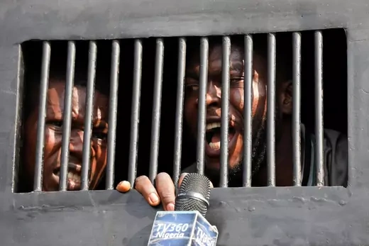 Two men look out from behind bars in a police vehicle. One speaks into a microphone, which reads "TV360 Nigeria," being held near his face.
