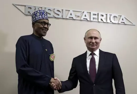 Nigerian President Muhammadu Buhari (left) and Russian President Vladimir Putin (right) shake hands. Buhari wears traditional Islamic dress, while Putin is wearing a jacket and tie. A metal sign reading "Russia-Africa" is seen behind the two.