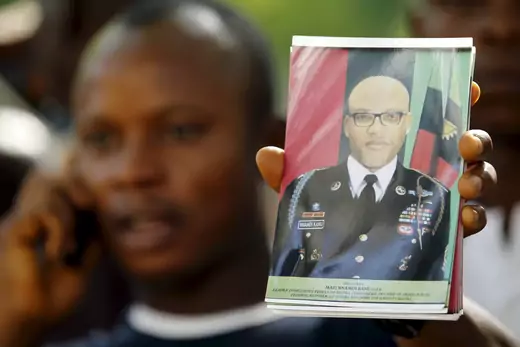 A man, out of focus in the back of the picture while speaking on a mobile phone, holds up a picture of Nnamdi Kanu, leader of the Indigenous People of Biafra, wearing an official military uniform.