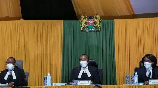 Three Kenyan judges, one sitting below the Kenyan coat of arms, are seen seated wearing judicial garb and medical facemasks.