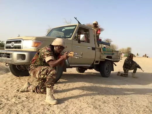Two solders crouch on one knee, one with an assault rifle pointed forward, the other with his rifle aimed at the ground. Behind them is a Toyota pickup truck with a mounted gun in the back, with another soldier manning the gun.