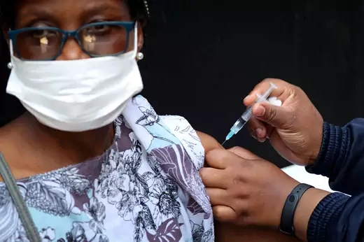 A woman, wearing glasses and a mask, has her sleeve rolled up as another person inserts a syringe in her upper arm to deliver a COVID-19 vaccination.