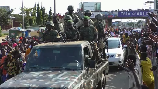 Several men in military uniform ride in the back of a military pickup through the streets. Supporters line the streets with their arms outstretched.