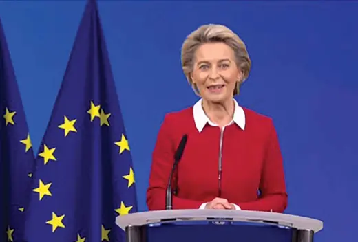 Ursula von der Leyen, wearing a red jacket, stands before a podium, with the European flag behind her.