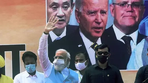 Modi waves while standing in front of a poster of Biden, Suga, and Morrison.