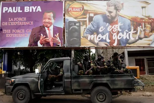 Soldiers brandishing rifles sit in the back of a camouflaged pickup truck. An election advertisement board of Cameroon President Paul Biya is seen behind them alongside another billboard with an advertisement.