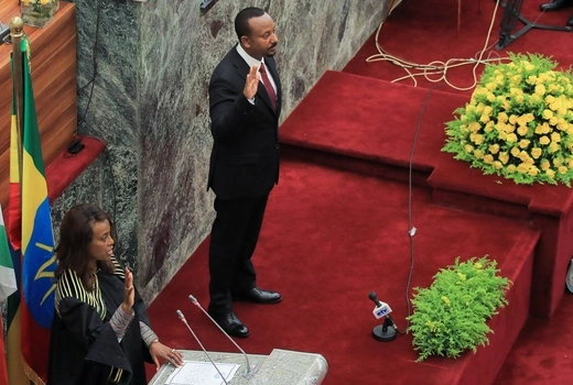 Ethiopian Prime Minister Abiy Ahmed stands and raises his right hand beside a woman standing at a podium with her hand raised. An Ethiopian flag sits behind both of them.