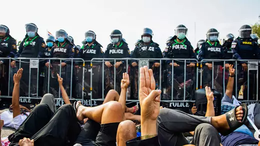 Protesters lay on the ground in front of a line of police and raise their hands in a three-finger salute.