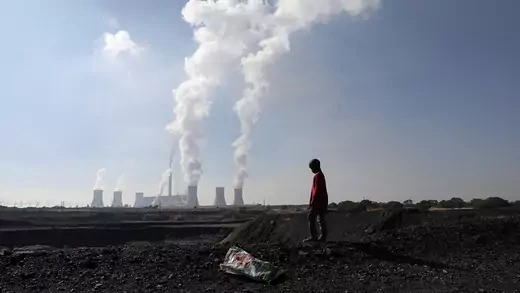 A person stands in front of coal-fired power plants billowing smoke into the sky.