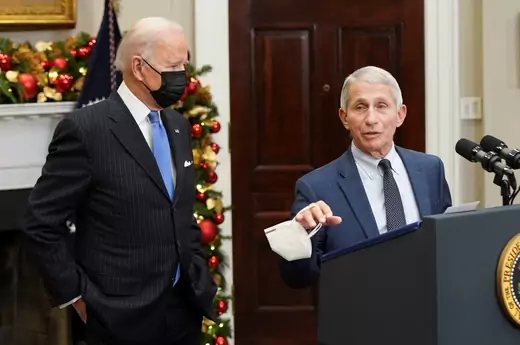 U.S. President Joe Biden stands wearing a mask behind Dr. Anthony Fauci, a top infectious disease expert, who is speaking at a podium with a mask in hand.