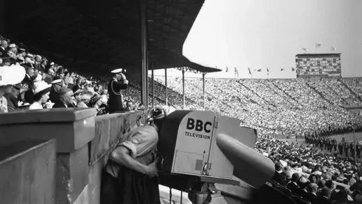 A BBC camera operator films the opening ceremony in a crowded stadium.